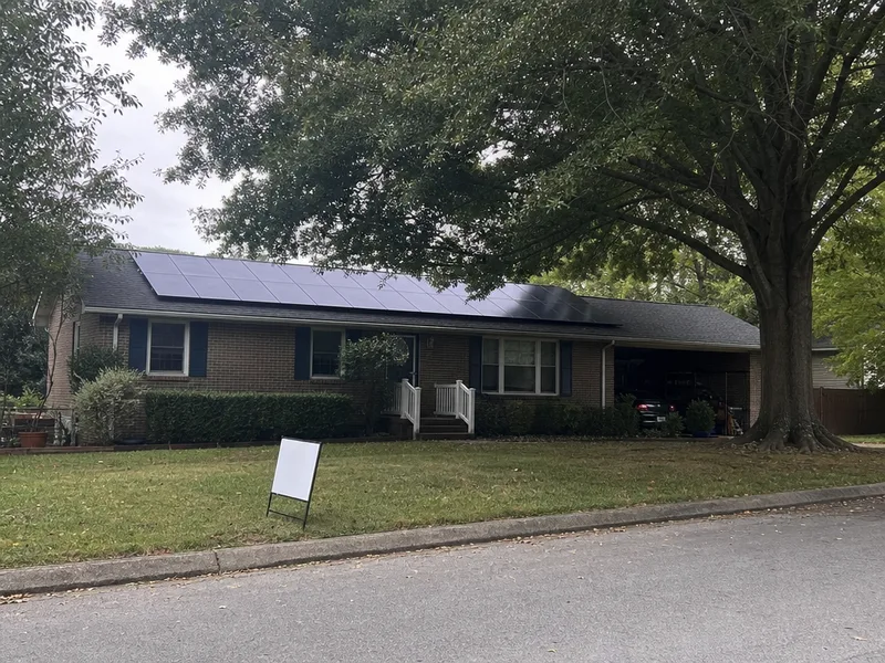 A single storey brick house with solar modules on its roof seen from across the street, a small unlettered yard sign near the kerb, a mature oak throwing shade across one end of the array