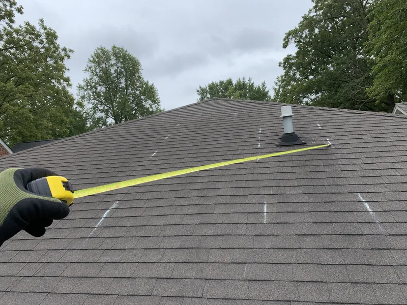 A gloved hand holding a tape measure across a bare asphalt shingle roof plane with chalk marks where the mounting rails will go, the ridge and a plumbing vent in frame