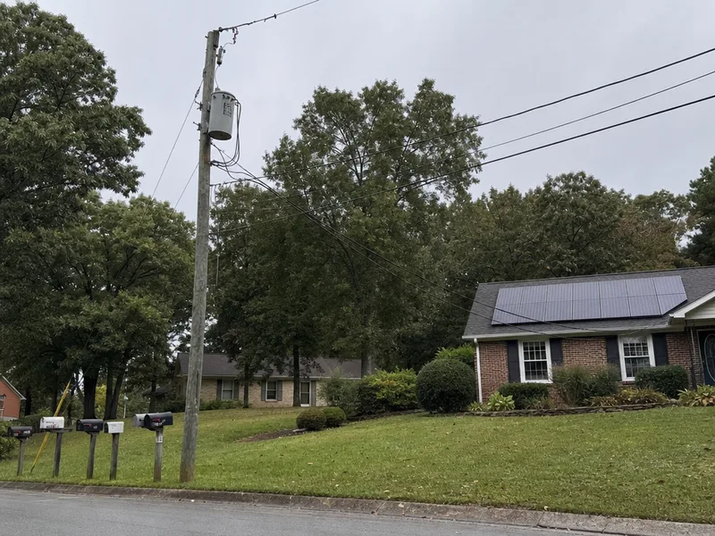 A pole mounted transformer on a wooden utility pole at the edge of a residential street, its overhead service drop running toward a brick house with solar modules on the roof