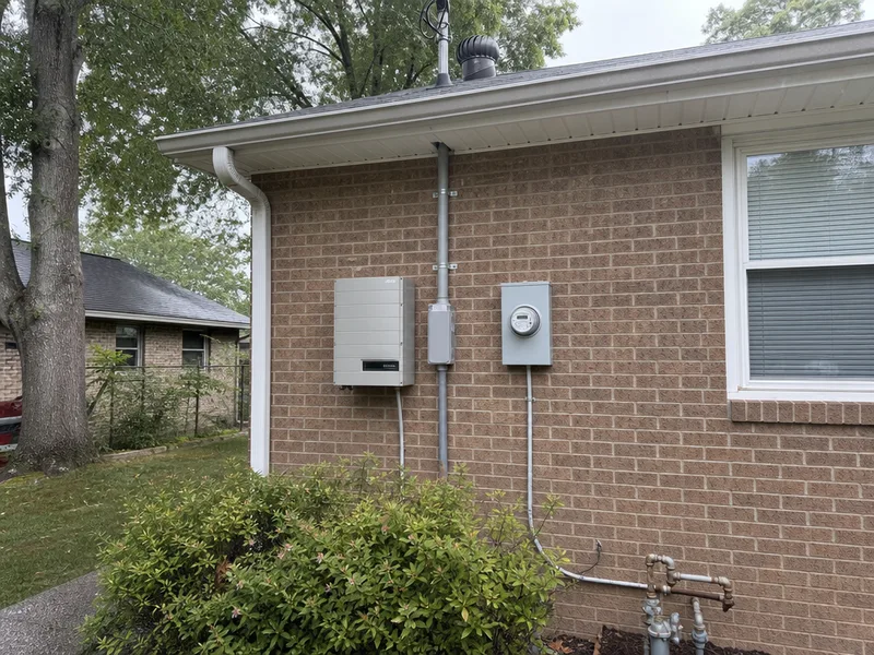 A grey string inverter, an AC disconnect and a utility meter mounted together on the brick wall of a Middle Tennessee house