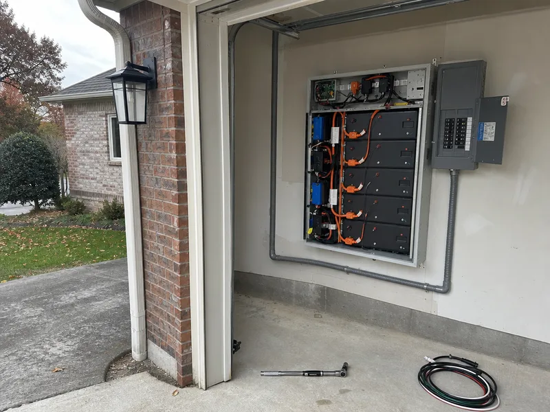A solar battery installation part way through in an attached garage, the cabinet cover off with its cell modules and orange DC cabling exposed, the main electrical panel beside it and a torque wrench on the floor