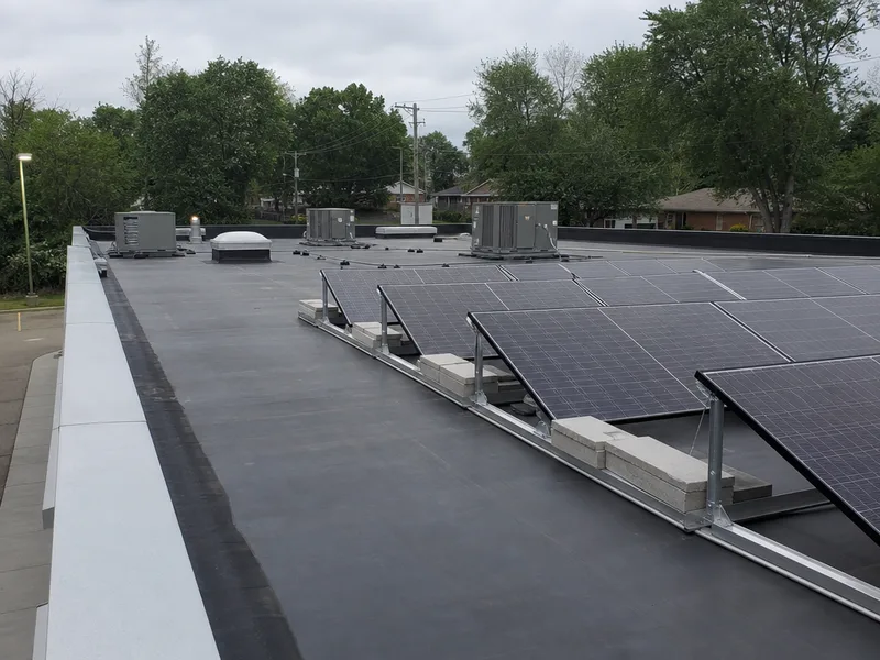 A commercial solar installation on a low slope membrane roof, tilted modules on ballasted trays weighted with concrete blocks, rooftop HVAC units and the parapet edge beyond